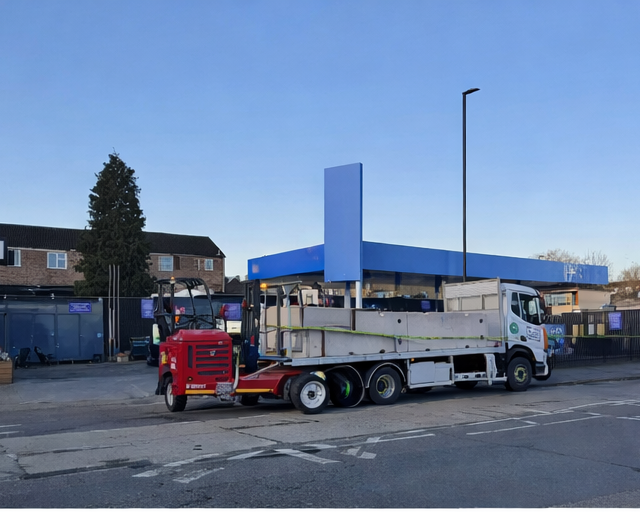 Flatbed truck unloading concrete barriers with mounted forklift on urban street near commercial site.