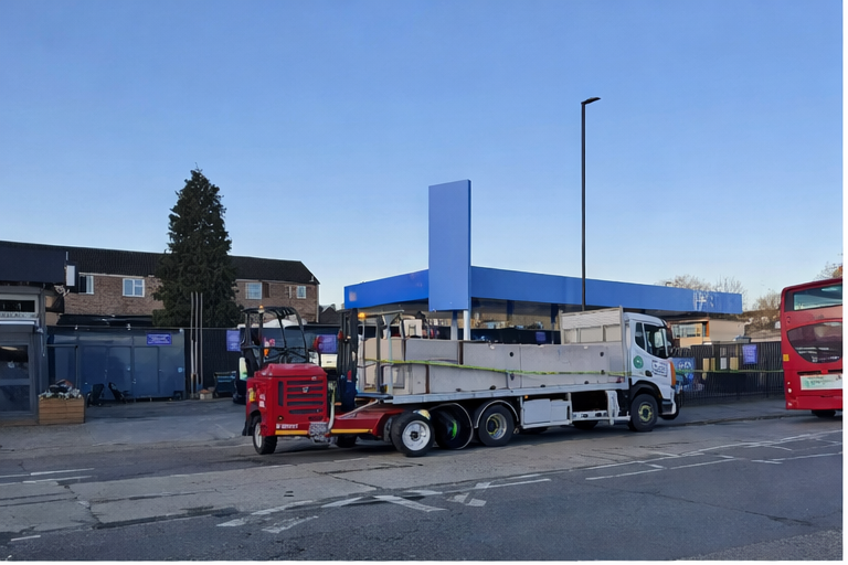 Flatbed truck unloading concrete barriers with mounted forklift on urban street near commercial site.