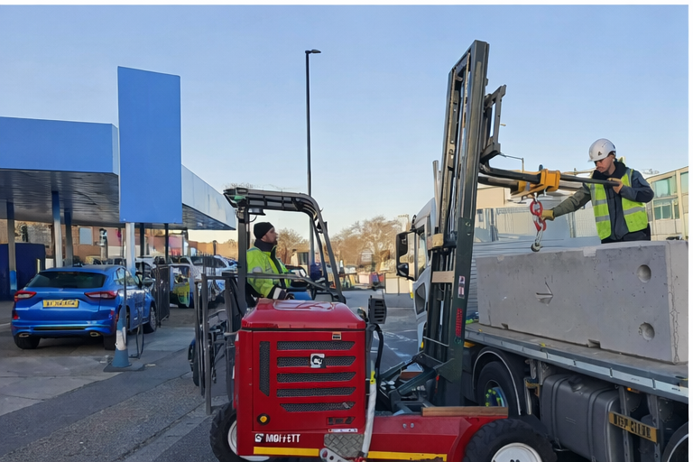 Flatbed truck unloading concrete barriers with mounted forklift on urban street near commercial site.